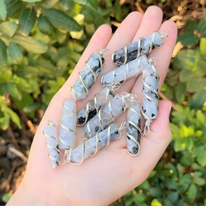 May include: A collection of gemstone pendants, each featuring a cylindrical stone wrapped in silver-colored wire. The stones display a mix of white, gray, and black patterns. The pendants are held in a hand against a backdrop of green foliage.