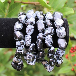 May include: Five black and white snowflake obsidian bracelets stacked on a black velvet display stand. The bracelets are made of tumbled stones with a speckled pattern.