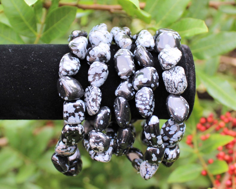 May include: Five black and white snowflake obsidian bracelets stacked on a black velvet display stand. The bracelets are made of tumbled stones with a speckled pattern.