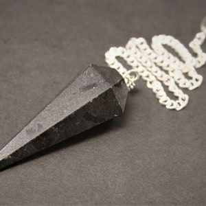 May include: A black, faceted pendulum with a silver chain and a clear crystal bead at the end. The pendulum is cone-shaped and appears to be made of a dark stone. The chain is linked and attached to the pendulum.