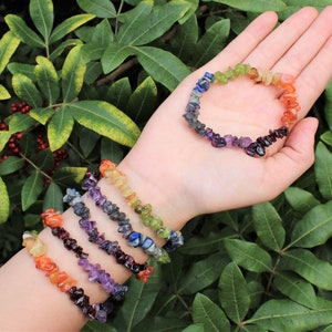 May include: Seven bracelets made of small, chipped gemstones in rainbow colors. The bracelets are stacked on a person's wrist.