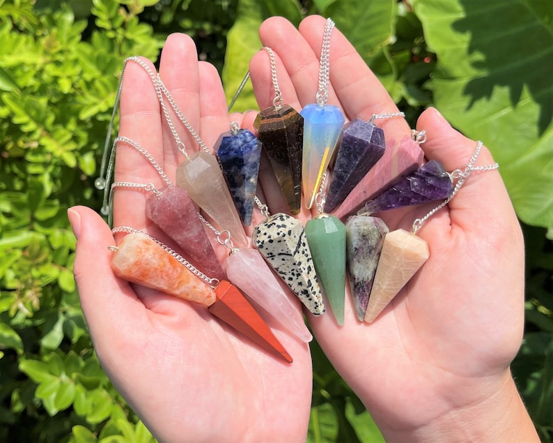 May include: A collection of 12 crystal point pendulums with silver chains. The pendulums are made of various crystals, including amethyst, rose quartz, clear quartz, tiger's eye, and carnelian.