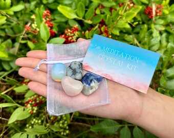 May include: A white organza bag filled with four tumbled crystals: blue, white, pink, and gray. The bag is in a hand, and a blue and pink card with the text "Meditation Crystal Kit" is visible.