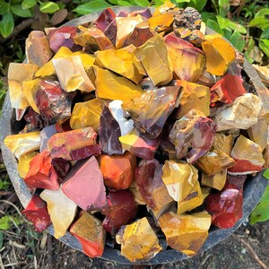 May include: A close-up shot of a dark gray stone bowl filled with raw, colorful rocks. The rocks are a mix of red, yellow, and brown hues. The rough-cut stones vary in size and shape, with a natural, earthy appearance. Green foliage surrounds the bowl.