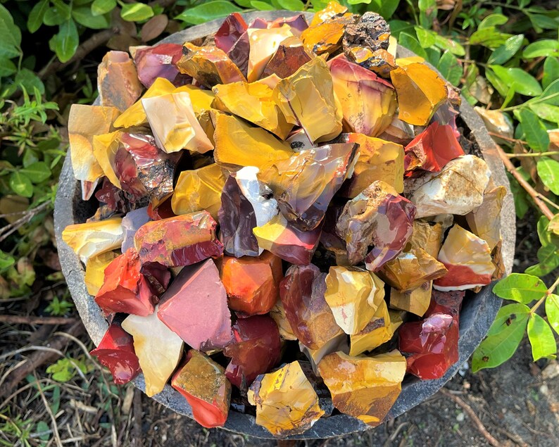 May include: A close-up shot of a dark gray stone bowl filled with raw, colorful rocks. The rocks are a mix of red, yellow, and brown hues. The rough-cut stones vary in size and shape, with a natural, earthy appearance. Green foliage surrounds the bowl.