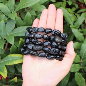 May include: A bracelet composed of multiple strands of polished, irregular black tourmaline stones. The stones are smooth, glossy, and dark, strung together to form a multi-layered accessory that wraps around the wrist area in a dense, textured formation.