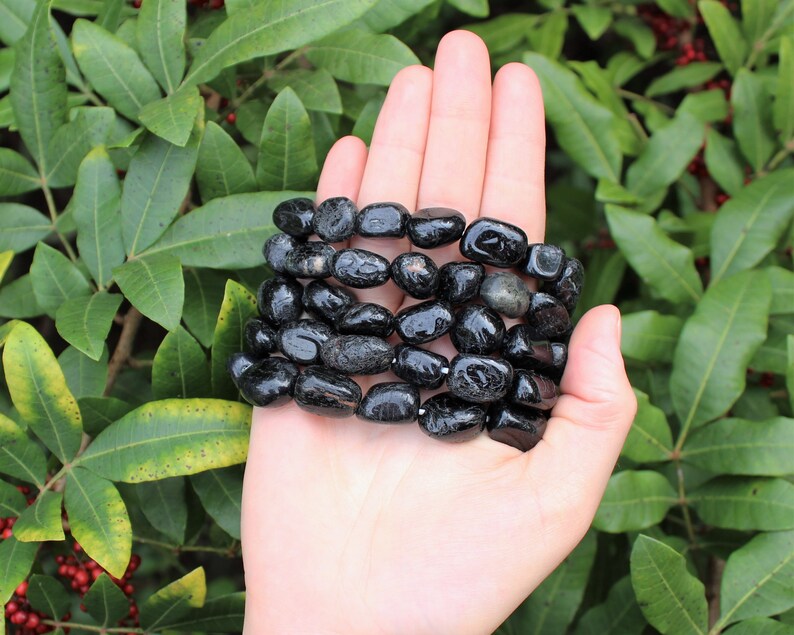 May include: A bracelet composed of multiple strands of polished, irregular black tourmaline stones. The stones are smooth, glossy, and dark, strung together to form a multi-layered accessory that wraps around the wrist area in a dense, textured formation.