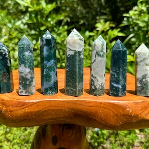 May include: Seven moss agate crystal towers of varying sizes and patterns are displayed on a wooden stand. The crystals feature green and white inclusions, with pointed tops. The stand is made of light brown wood, and the background is blurred green foliage.