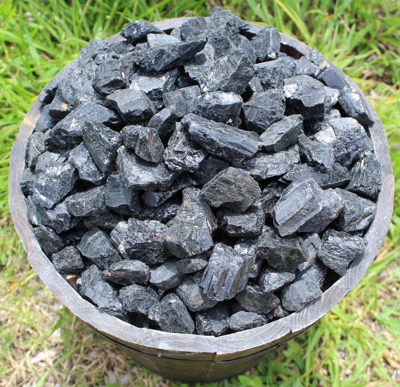 May include: A wooden bowl filled with black tourmaline rough crystals. The crystals are various sizes and shapes, with some having a pointed or elongated shape.
