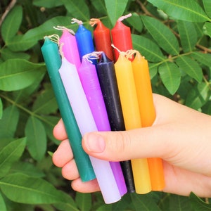 May include: A collection of ten thin, colorful candles in various shades of green, blue, red, purple, white, black, and yellow. The candles are held in a hand.