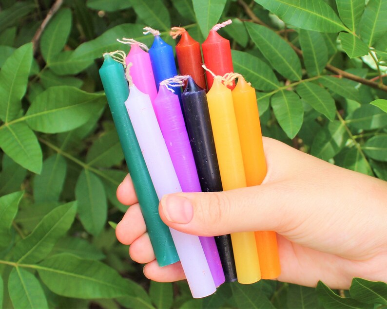 May include: A collection of ten thin, colorful candles in various shades of green, blue, red, purple, white, black, and yellow. The candles are held in a hand.