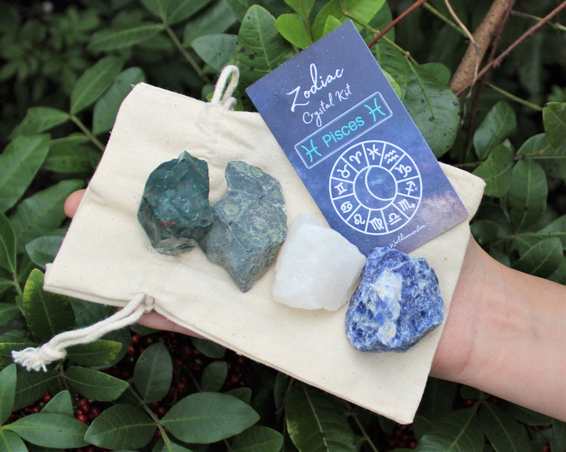 K&ouml;nnte beinhalten: Eine kleine wei&szlig;e Kordeltasche mit vier Kristallen darin. Die Kristalle sind gr&uuml;n, wei&szlig; und blau. Eine Karte mit dem Text "Zodiac Crystal Kit Pisces" liegt auf der Tasche.