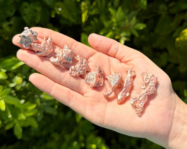 May include: A collection of eight small, irregular copper-colored metal nuggets with a textured, porous surface. The pieces vary in shape and size, displaying a metallic sheen and a reddish-brown hue, arranged in a line across the palm of a hand.