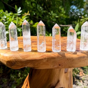 May include: Seven clear, hexagonal quartz crystal points arranged in a row on a polished, light brown wooden surface. Each crystal features a pointed termination and a transparent, multifaceted body with visible internal inclusions and fractures.