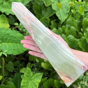 May include: A tall, polished stone tower with a smooth, light green surface featuring translucent bands and subtle orange-brown mineral inclusions. The object has a pointed, faceted top and a flat, rectangular base, showcasing natural stone patterns.