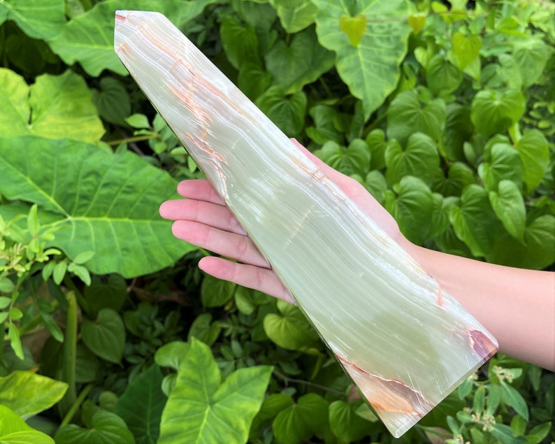 May include: A tall, polished stone tower with a smooth, light green surface featuring translucent bands and subtle orange-brown mineral inclusions. The object has a pointed, faceted top and a flat, rectangular base, showcasing natural stone patterns.