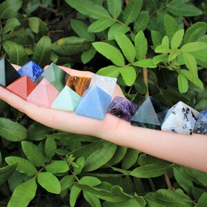 Puede incluir: Una colecci&oacute;n de pir&aacute;mides de cristal de colores, incluyendo verde, rojo, rosa y azul. Las pir&aacute;mides se muestran en una mano sobre un fondo de follaje verde. Cada pir&aacute;mide tiene una superficie lisa y pulida.