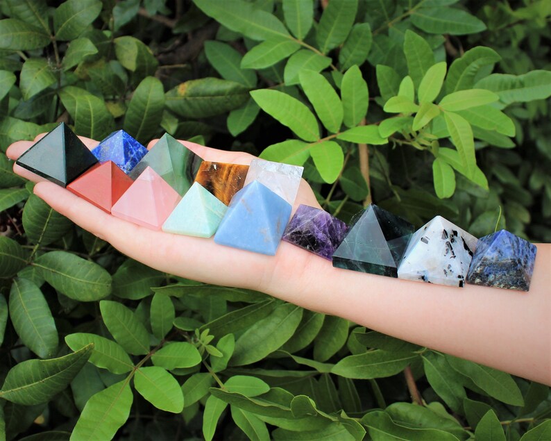 Puede incluir: Una colecci&oacute;n de pir&aacute;mides de cristal de colores, incluyendo verde, rojo, rosa y azul. Las pir&aacute;mides se muestran en una mano sobre un fondo de follaje verde. Cada pir&aacute;mide tiene una superficie lisa y pulida.