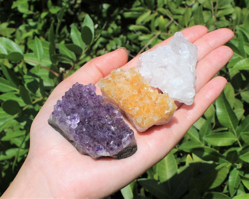 May include: Three raw crystal clusters in a hand. The clusters are amethyst, citrine, and clear quartz. The amethyst is purple, the citrine is yellow, and the quartz is clear.