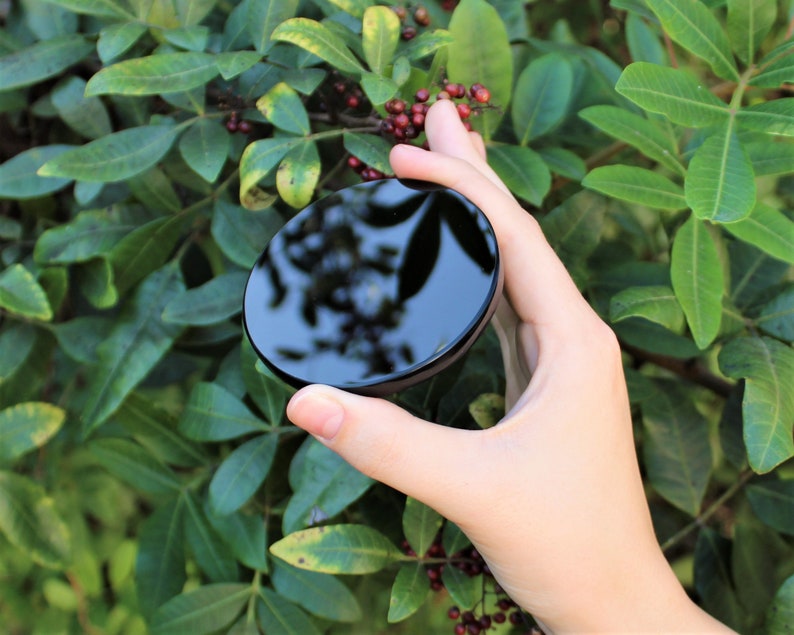 May include: A black round mirror with a reflective surface. The mirror is being held in a hand.