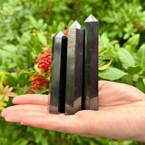 Pu&ograve; includere: Quattro punte di cristallo di ossidiana nera, anche note come vetro nero, sono disposte in fila nella mano di una persona. Le punte sono lisce e lucidate, con un leggero bagliore.
