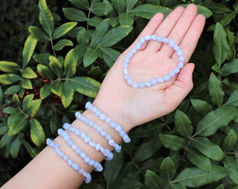 May include: A collection of six stretch bracelets made from light blue, translucent, tumbled gemstone beads. One bracelet is held in the palm of the hand, while five additional bracelets are stacked around the wrist, showcasing the smooth texture.