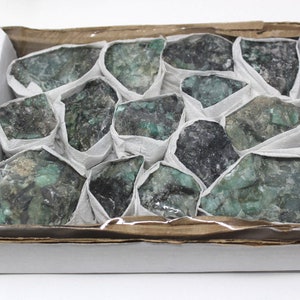 May include: A box filled with rough, green emerald stones. The stones are wrapped in white tissue paper and are arranged in a single layer.