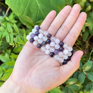 May include: A bracelet made of amethyst, rose quartz, and clear quartz beads. The bracelet is layered and has a purple, pink, and white color scheme.