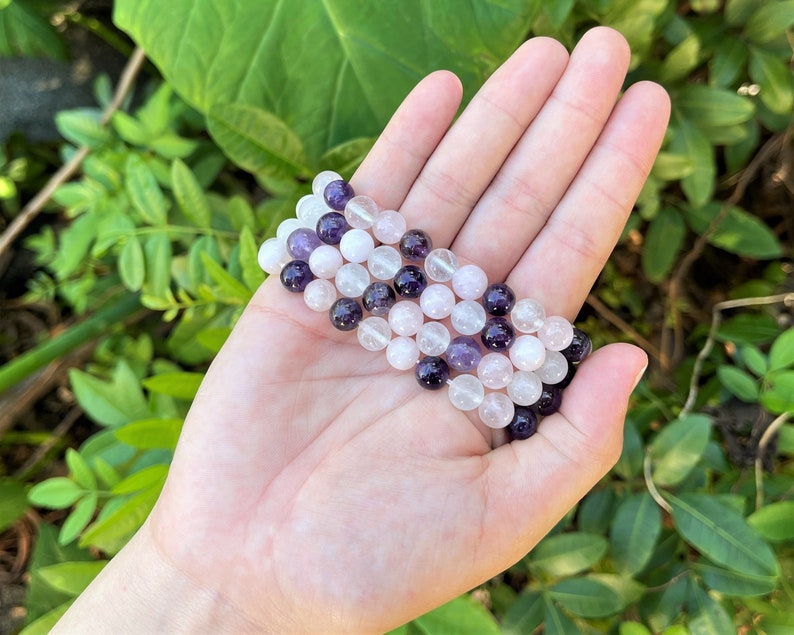 May include: A bracelet made of amethyst, rose quartz, and clear quartz beads. The bracelet is layered and has a purple, pink, and white color scheme.