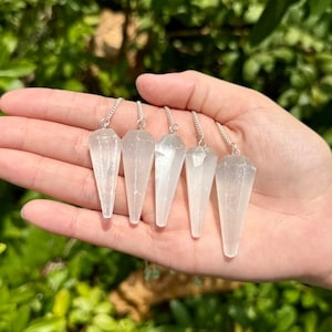May include: Five white selenite crystal pendulums with silver chains. The pendulums are all the same size and shape, and they are hanging from the chains.
