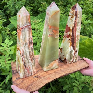 May include: Three polished stone obelisks with pointed tops, featuring banded patterns in shades of translucent green, cream, and deep reddish-brown, displayed on a natural-edged wooden board with a visible wood grain and dark knots.