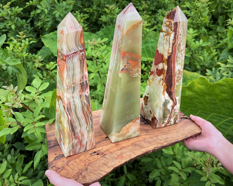 May include: Three polished stone obelisks with pointed tops, featuring banded patterns in shades of translucent green, cream, and deep reddish-brown, displayed on a natural-edged wooden board with a visible wood grain and dark knots.