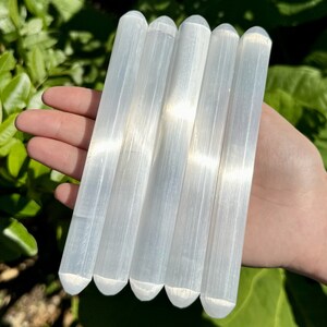 May include: Five white selenite wands, also known as selenite sticks, are arranged in a row. The wands are smooth and have a natural, striated texture.