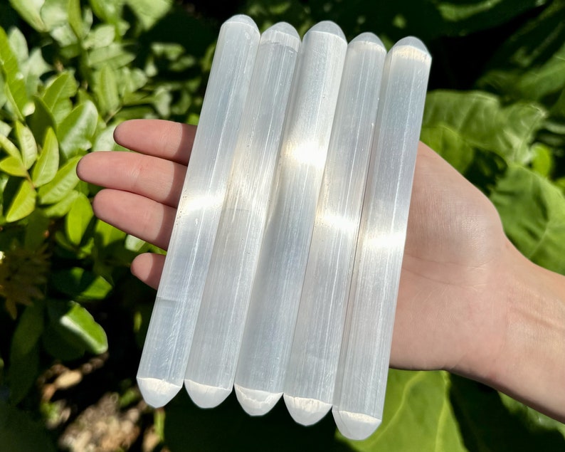 May include: Five white selenite wands, also known as selenite sticks, are arranged in a row. The wands are smooth and have a natural, striated texture.