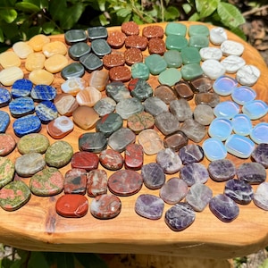 May include: A collection of polished gemstones in various colors, including yellow, blue, green, red, and purple, arranged on a wooden surface. The stones vary in shape and size, with a polished, smooth texture.