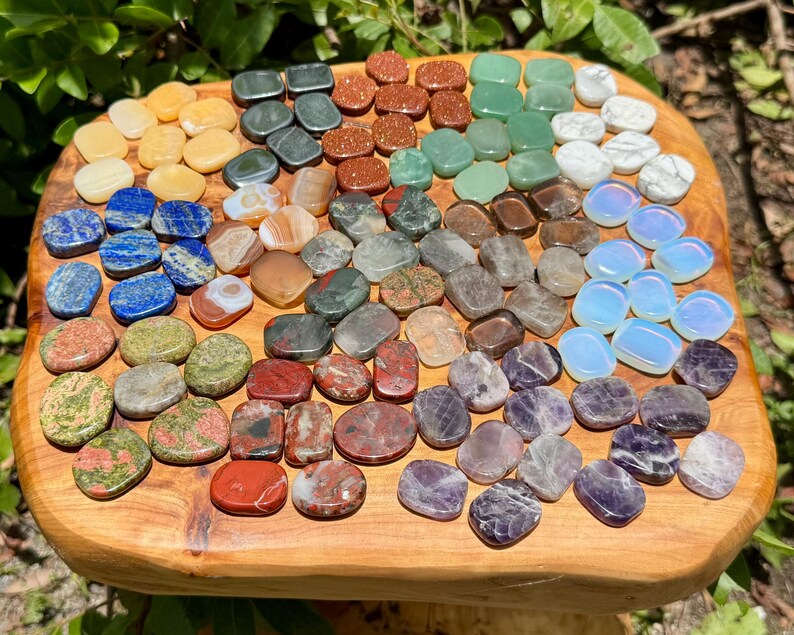 May include: A collection of polished gemstones in various colors, including yellow, blue, green, red, and purple, arranged on a wooden surface. The stones vary in shape and size, with a polished, smooth texture.