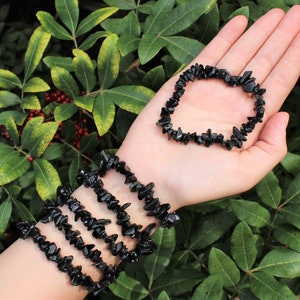 May include: Four black chip gemstone bracelets stacked on a wrist, with a fifth bracelet lying on a hand in front of a green leafy background.