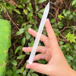 May include: A white selenite wand with a pointed tip. The wand is smooth and polished, and it is held in a hand against a green leafy background.