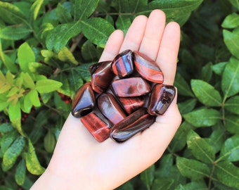 May include: A collection of red tiger's eye tumbled stones. The stones are smooth and polished, with a reddish-brown color and a golden sheen.