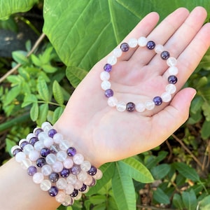 May include: Two bracelets made of amethyst and clear quartz beads. The bracelets are stacked on a person's wrist. The bracelets are a light purple and white color.