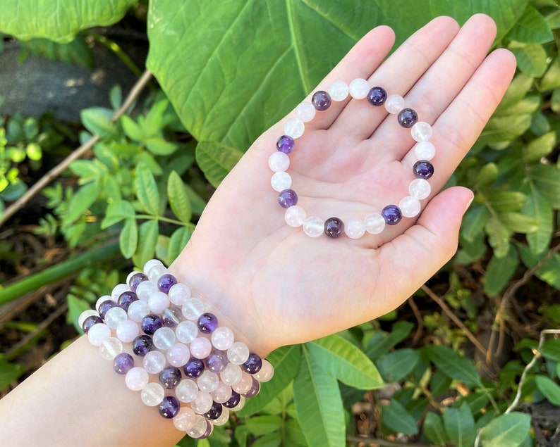 May include: Two bracelets made of amethyst and clear quartz beads. The bracelets are stacked on a person's wrist. The bracelets are a light purple and white color.