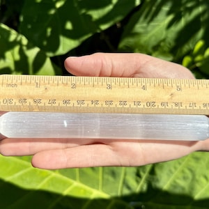 May include: A white selenite wand measuring approximately 5 inches long. The wand is smooth and has a slightly rounded end. It is being held in a hand for size reference.