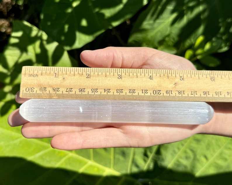 May include: A white selenite wand measuring approximately 5 inches long. The wand is smooth and has a slightly rounded end. It is being held in a hand for size reference.