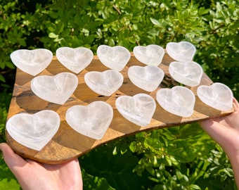 May include: A collection of heart-shaped, translucent white crystal bowls arranged on a wooden serving board. The bowls are held by a person, with a backdrop of green foliage. The bowls are approximately 4 inches wide.