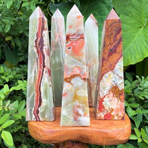 May include: Five polished stone obelisks with pointed tops, featuring banded patterns in shades of green, red, orange, and white. The mineral towers display natural veining and translucent layers, standing upright on a smooth, rounded wooden base.