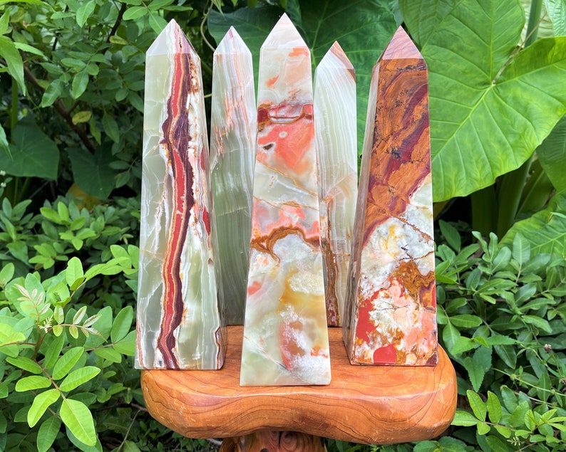 May include: Five polished stone obelisks with pointed tops, featuring banded patterns in shades of green, red, orange, and white. The mineral towers display natural veining and translucent layers, standing upright on a smooth, rounded wooden base.