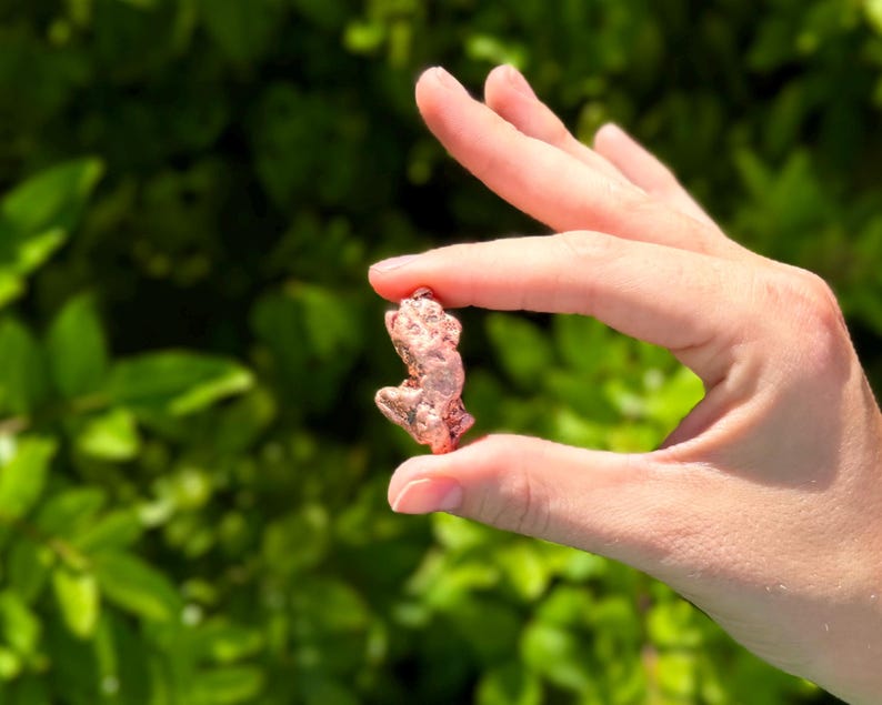 May include: A small, irregular copper nugget with a metallic, reddish-brown finish and a textured, uneven surface. The nugget displays a natural, raw appearance with various indentations and protrusions across its compact, solid form.