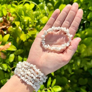 May include: White stone chip bracelets. One bracelet is held in a hand, while another is worn on the wrist. The bracelets are made of small, irregularly shaped white stones strung together. The background is a lush green bush.