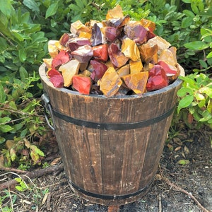 May include: A wooden bucket filled with a variety of colorful rough gemstones. The gemstones are in shades of red, yellow, and brown. The bucket has metal bands and a metal handle. The background is green foliage.