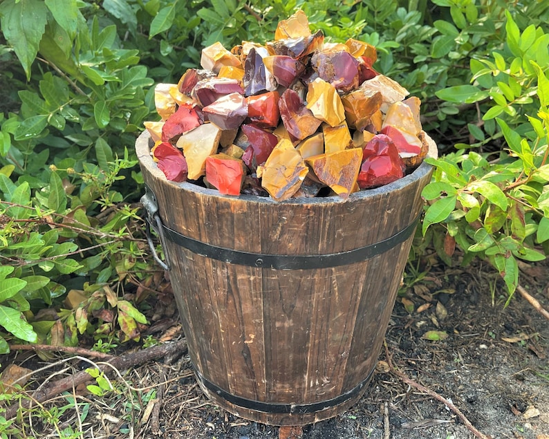 May include: A wooden bucket filled with a variety of colorful rough gemstones. The gemstones are in shades of red, yellow, and brown. The bucket has metal bands and a metal handle. The background is green foliage.
